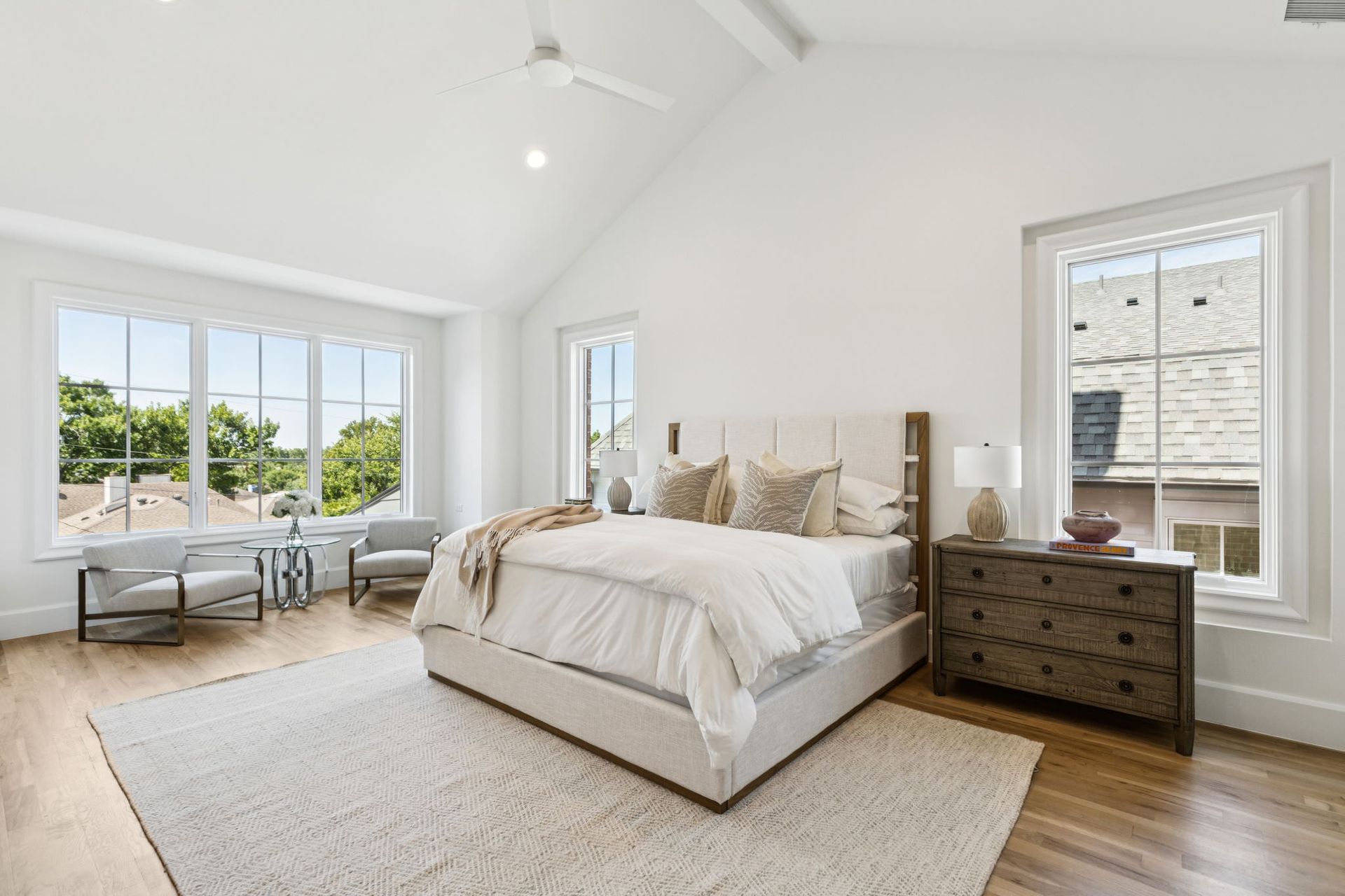Bright white bedroom with large windows, light wood floor, and neutral-toned furniture; bed, chairs, and dresser.