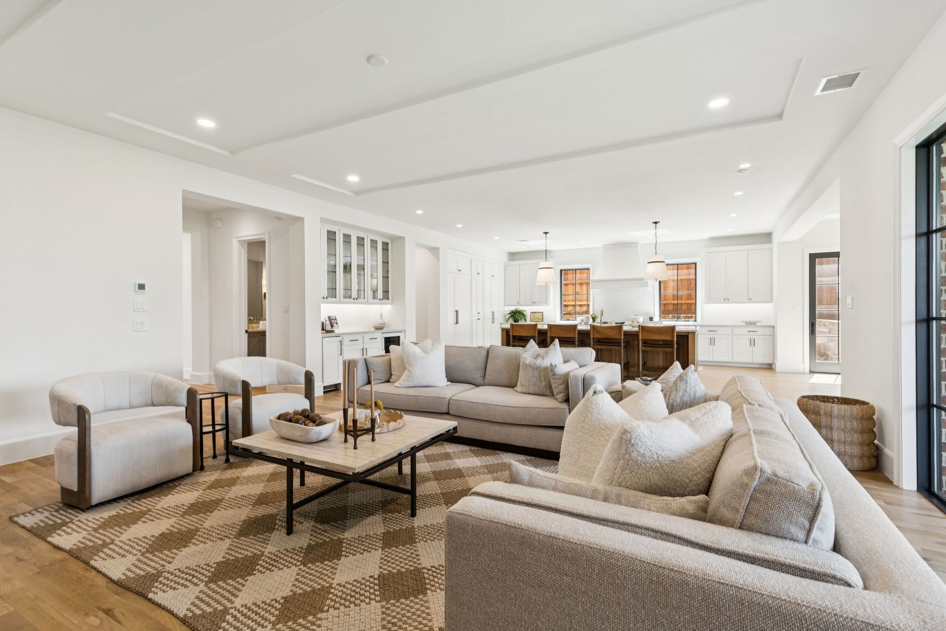 Spacious, light-filled living room with neutral-toned furniture, area rug, and open concept kitchen in the background.