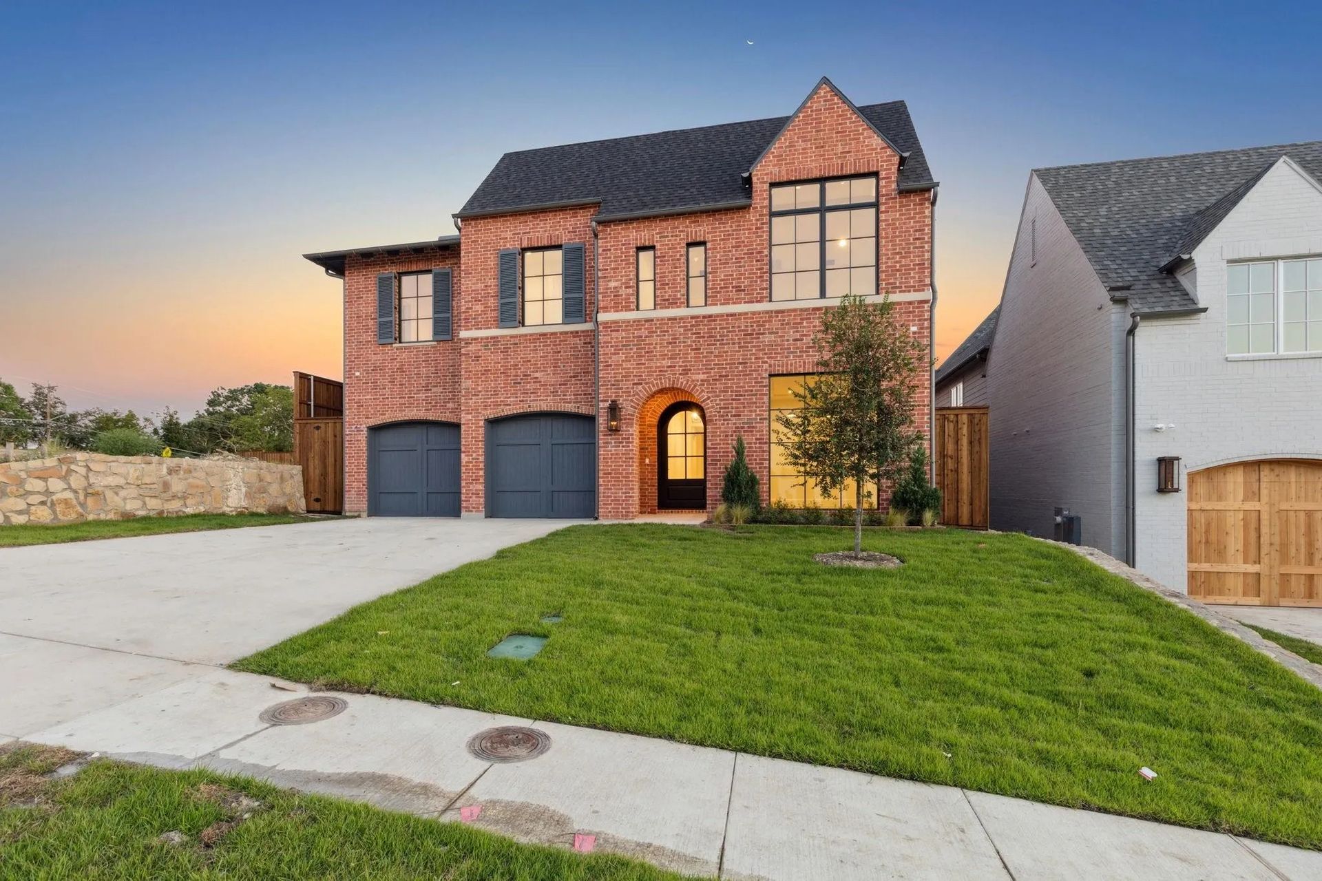 Two-story brick house with gray garage doors and a lawn. Next to a white house with wooden garage doors.