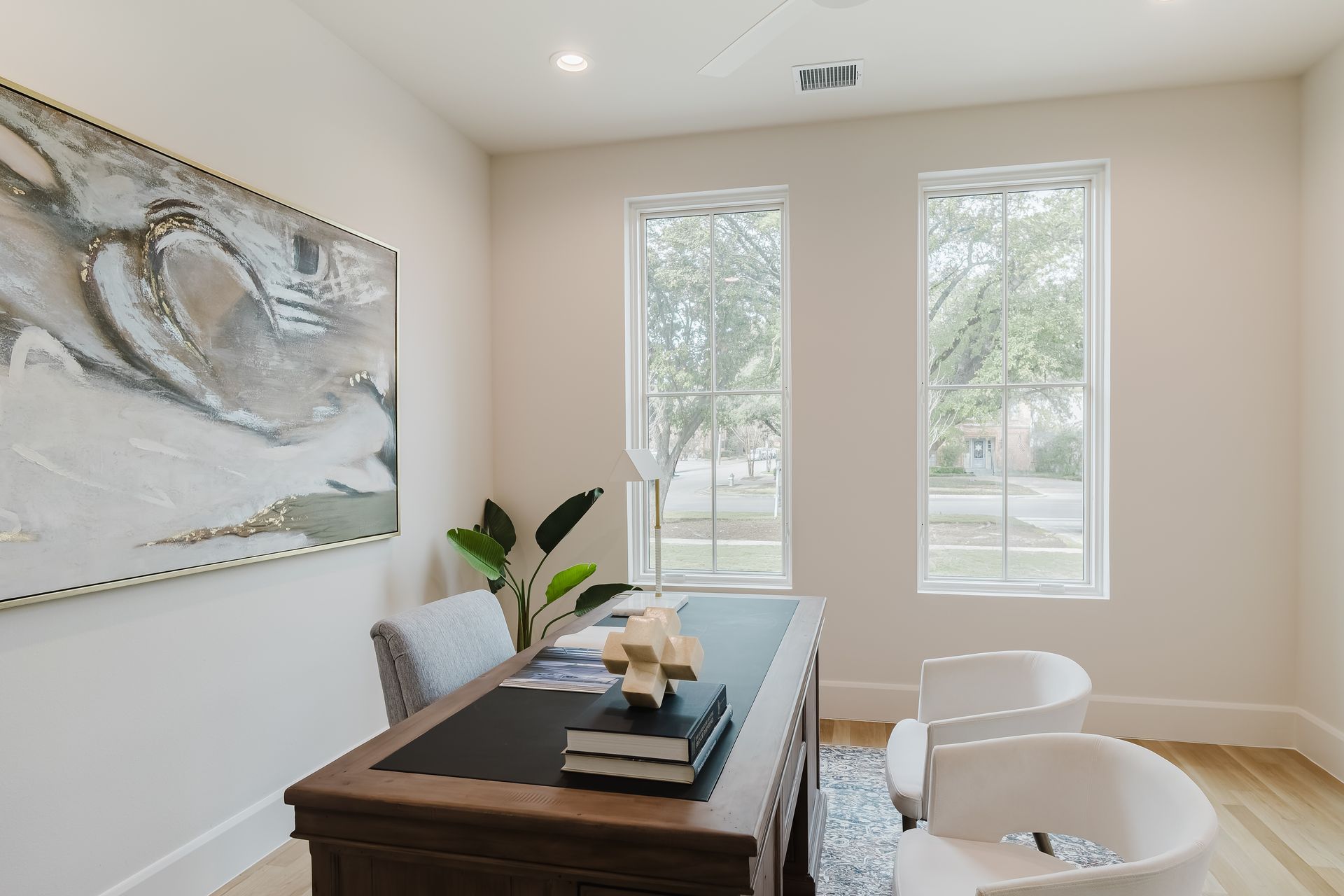 Office with desk, art, two windows with outside view, and two white chairs.