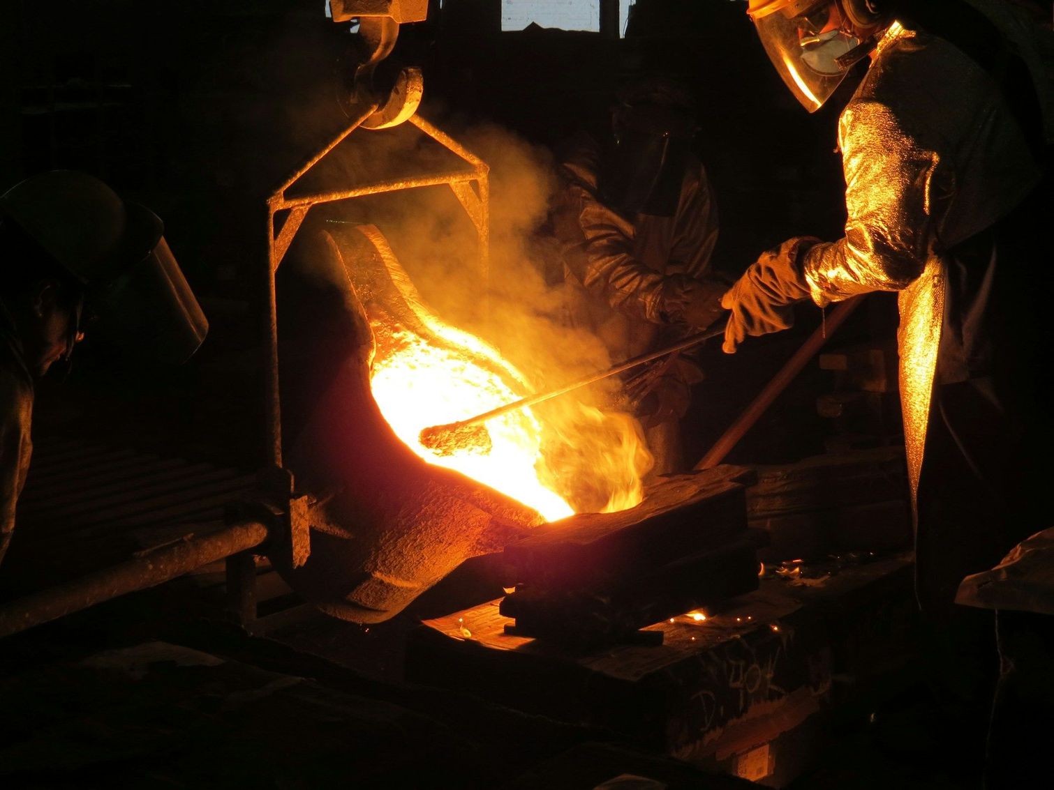 In una fonderia buia, gli operai versano metallo fuso in uno stampo, mentre fiamme arancioni brillanti ardono.