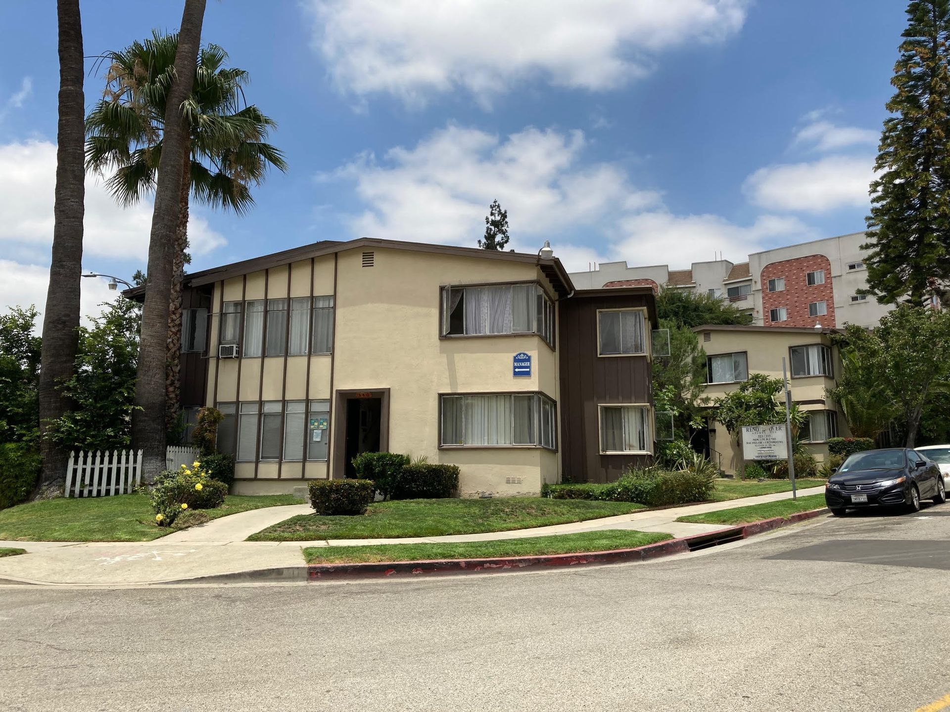 Apartment building on a corner lot; tan and brown exterior, palm trees, sunny day.