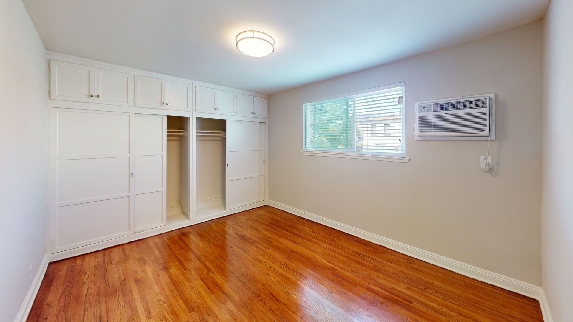 Empty bedroom with hardwood floors, white built-in closets, window, and air conditioner.