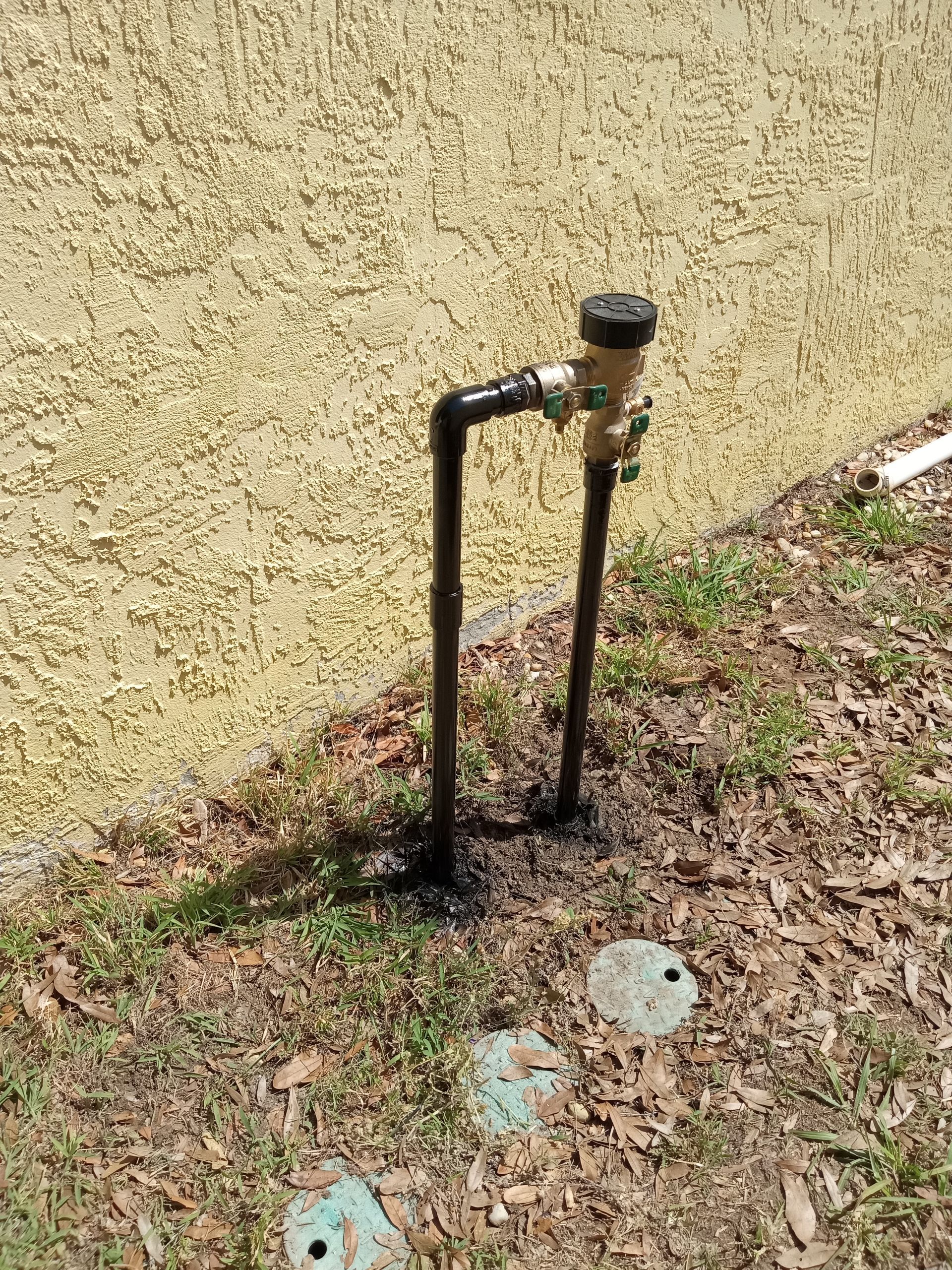 Black irrigation pipe and valve against a beige stucco wall beside patchy grass.