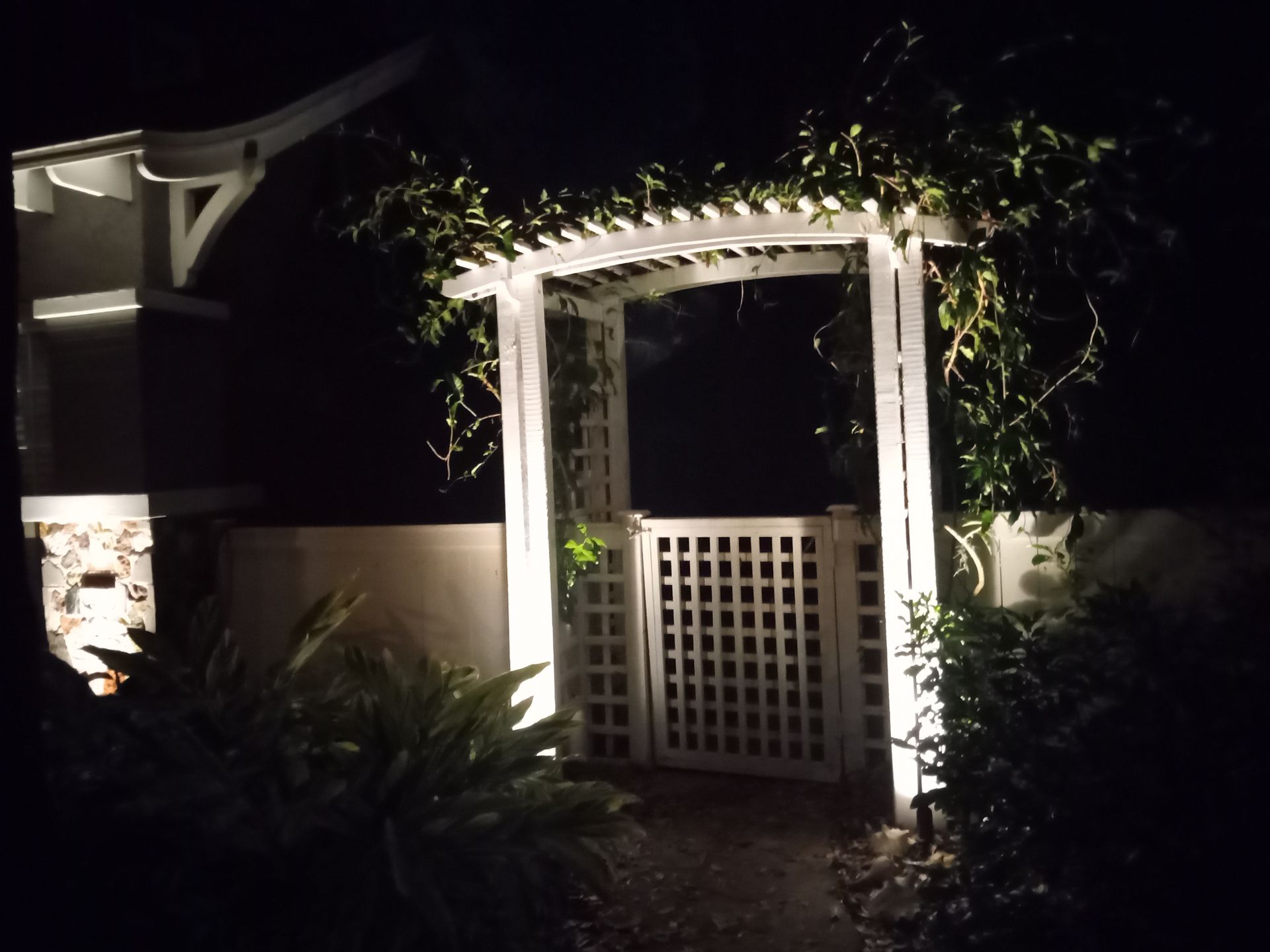 White garden arbor lit at night, framed by greenery and a small gate entrance