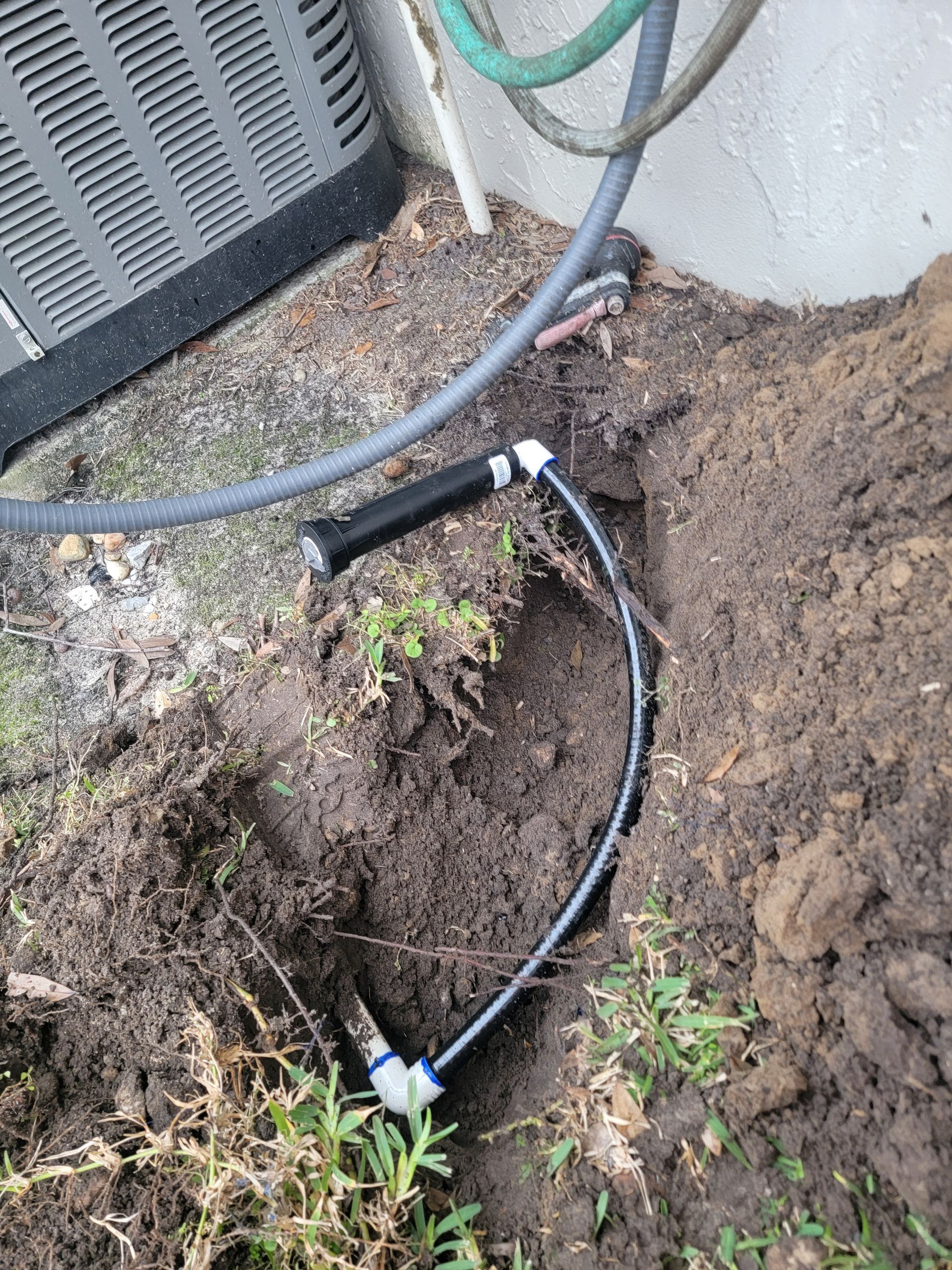 Black hose and wiring in a dirt trench beside an outdoor HVAC unit and wall.