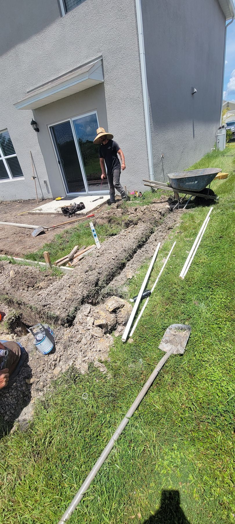 Worker trenching beside a gray house, with white pipes and tools on a grassy lawn.