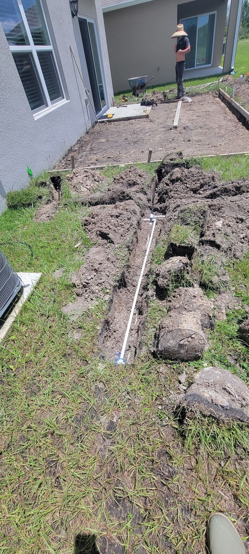 Trenched yard beside a house, with exposed pipes, dirt piles, and a worker in the background.