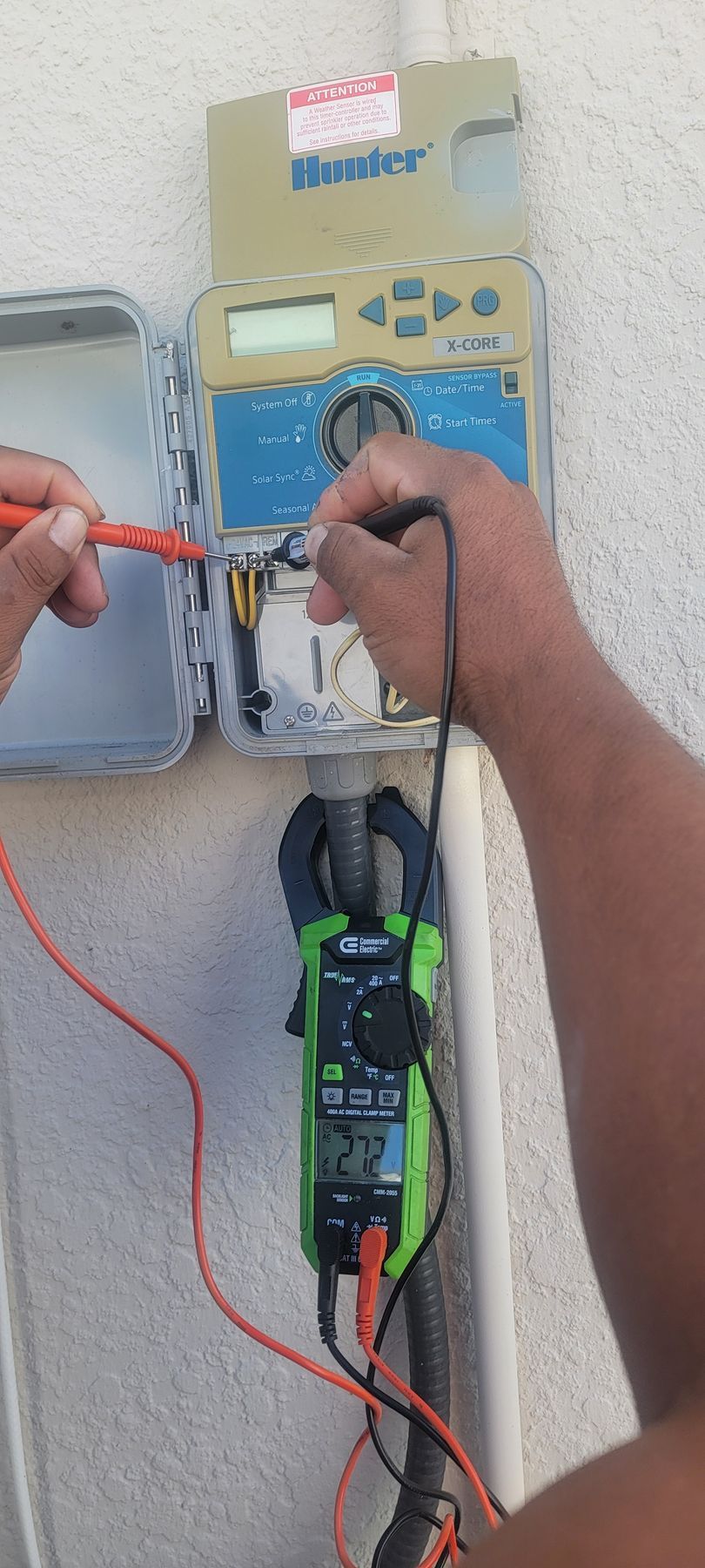 Person using test probes on an electrical meter panel with a clamp meter below.
