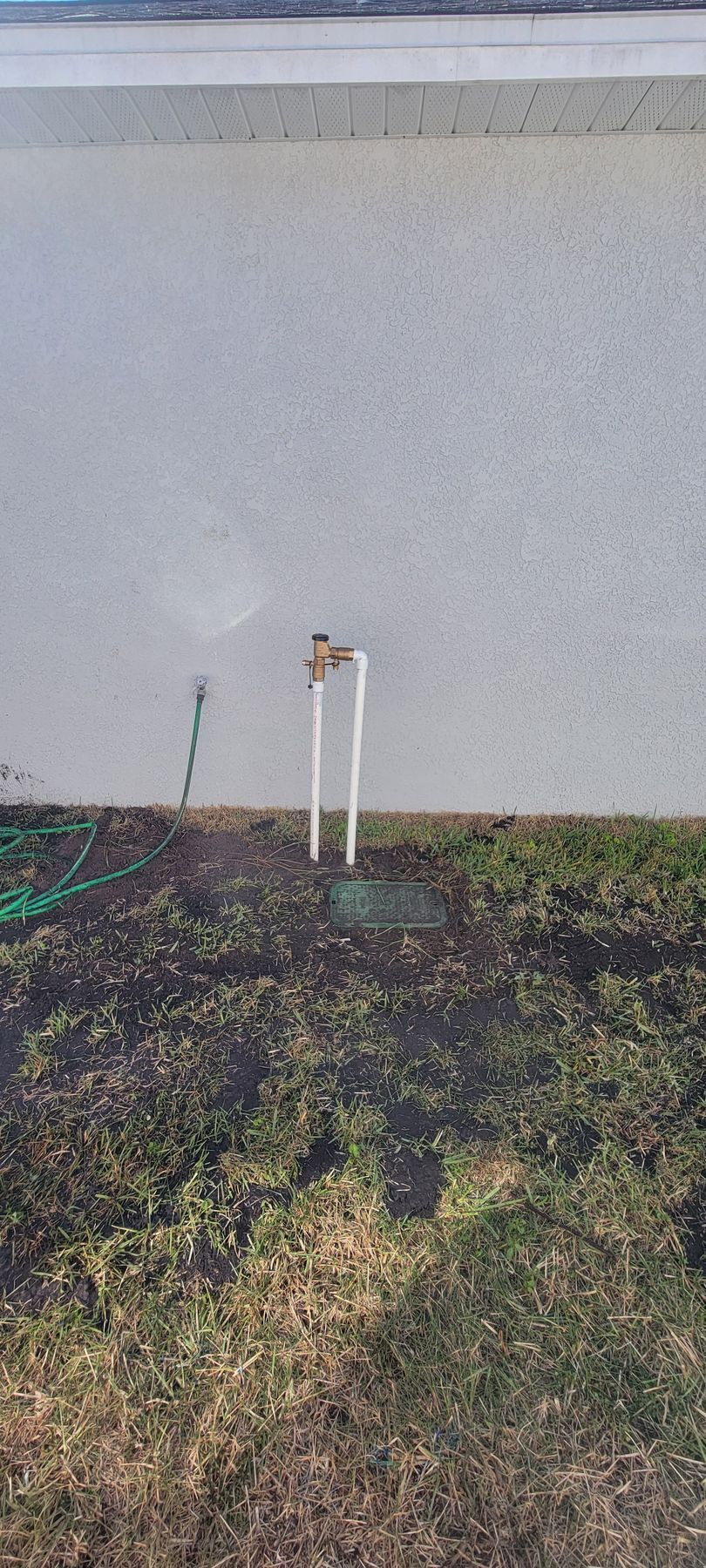 Two white pipes with a small valve against a pale wall above patchy grass and dirt.