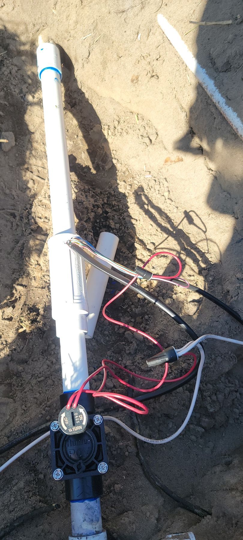 White PVC pipe with black and red wires and a small device, lying on dirt outdoors.