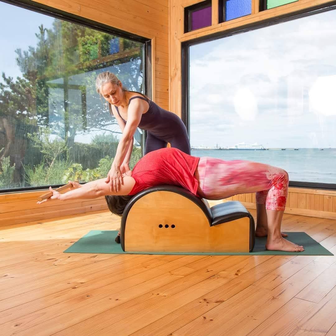 Mujer haciendo pilates en un barril con instructor, vista al mar de fondo.