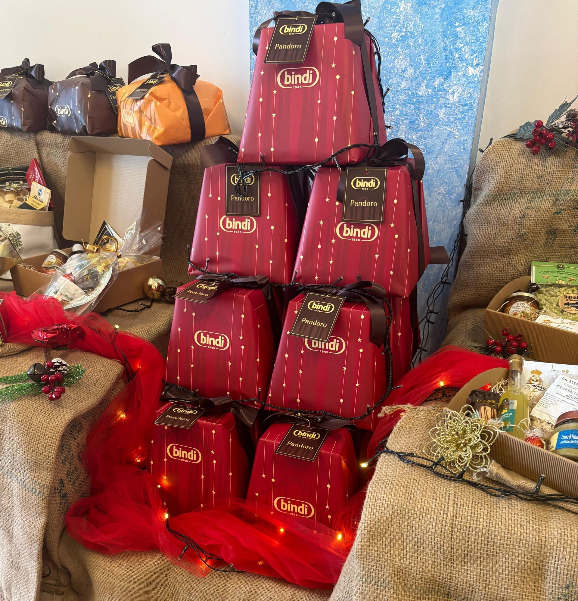 Un'esposizione di scatole di panettone del marchio Bindi disposte a piramide, avvolte in carta rossa con nastro e circondate da decorazioni in juta.