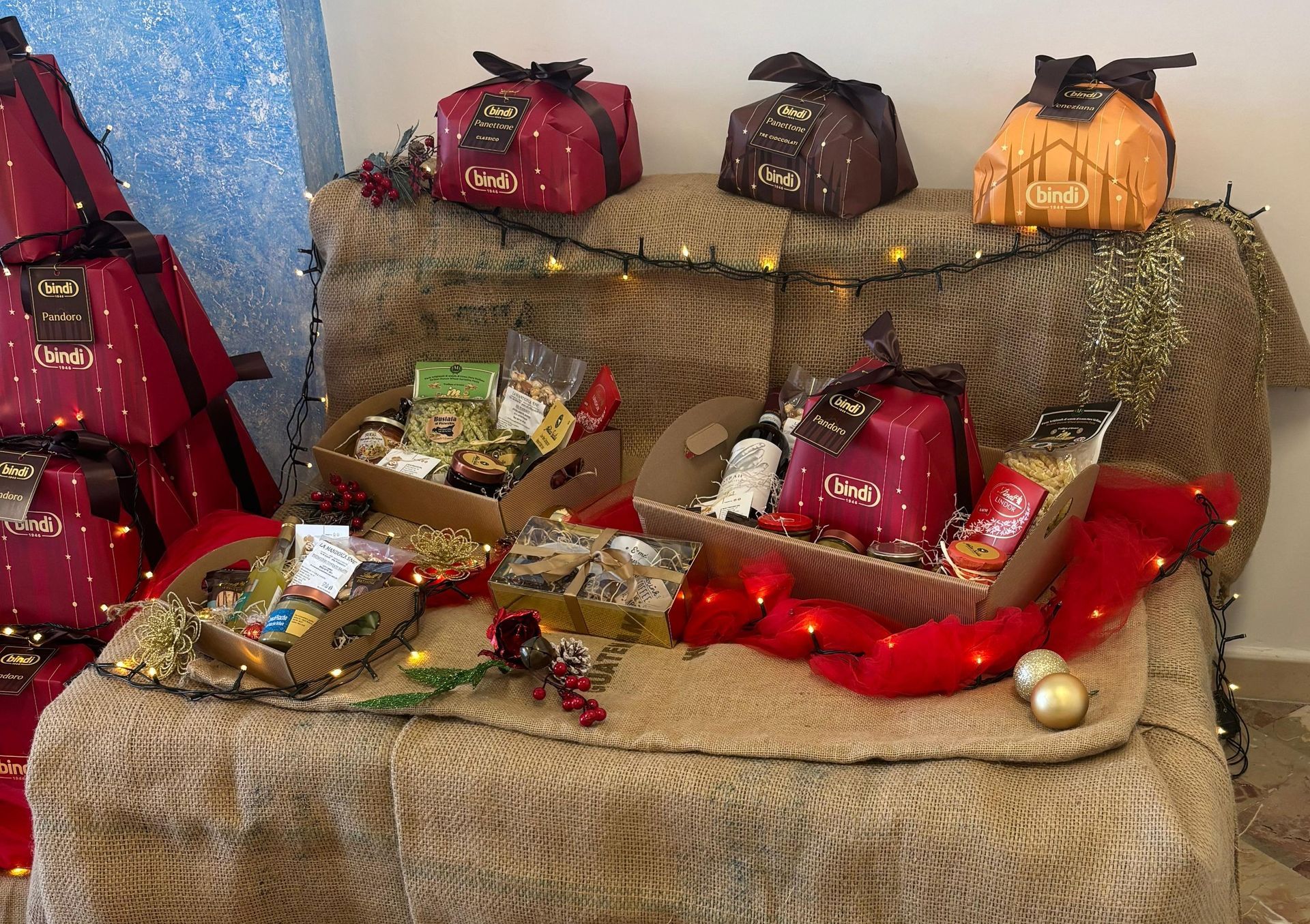Un'esposizione di cesti regalo, torte decorate in confezioni festive e ornamenti dorati su un tavolo ricoperto di tela di iuta.