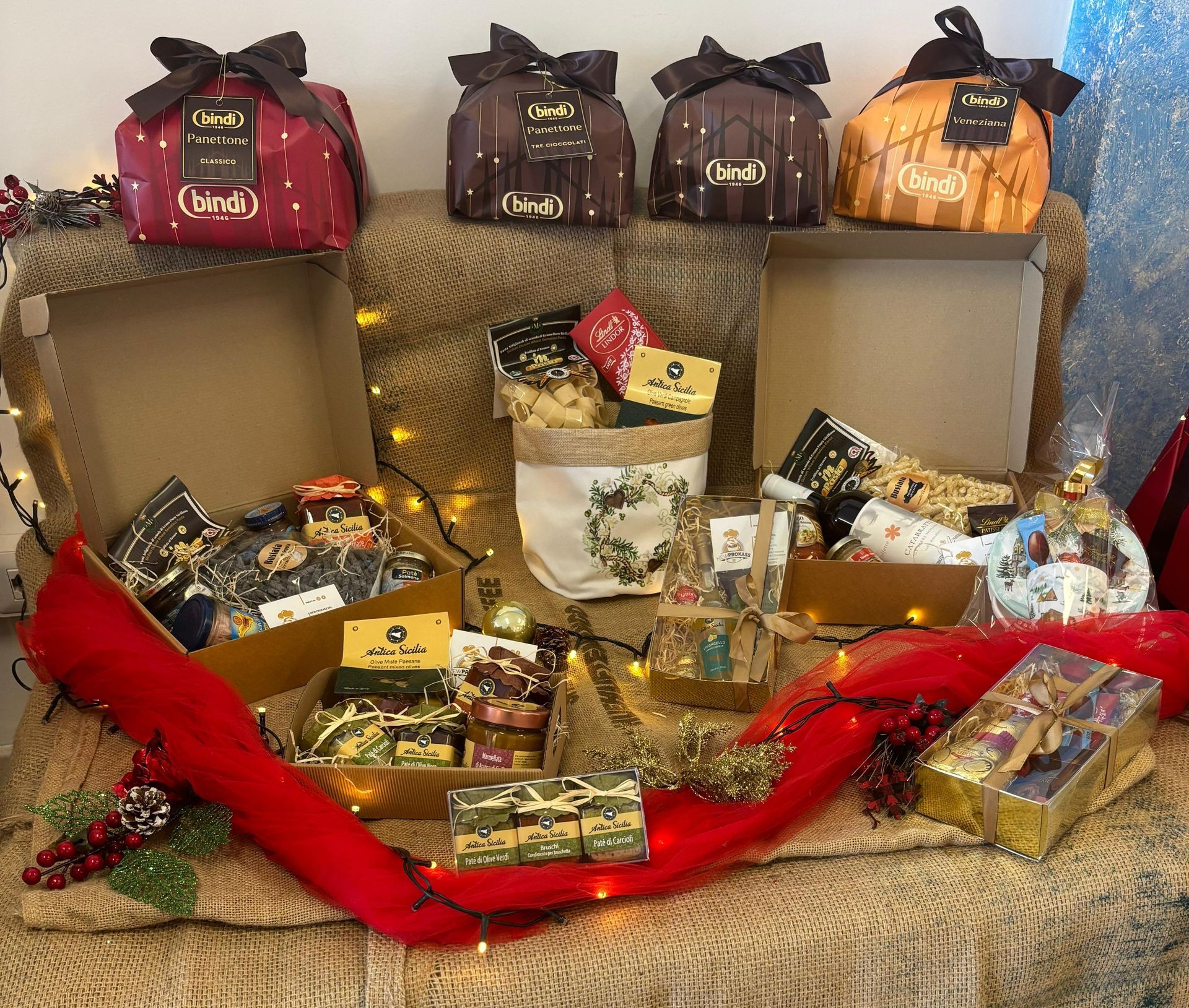 Una festosa collezione di cesti regalo, scatole decorative e prodotti alimentari a tema natalizio esposti su una superficie drappeggiata.