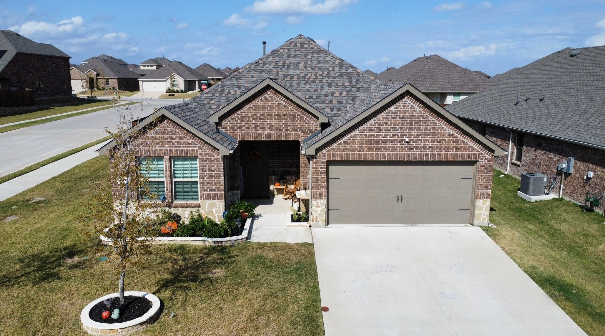A one-story suburban brick house with a gray shingled roof, a two-car garage, and a concrete driveway on a sunny day.