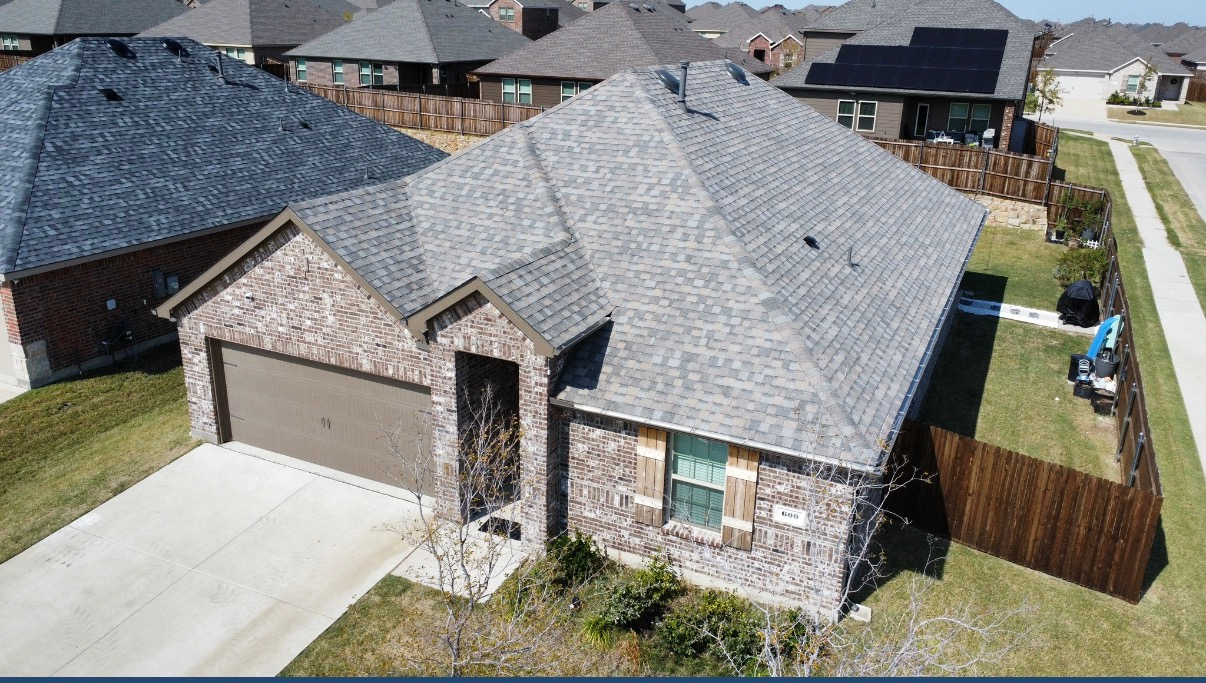 An aerial view of a suburban single-story brick house with a gray shingled roof, a garage, and a wooden fence.