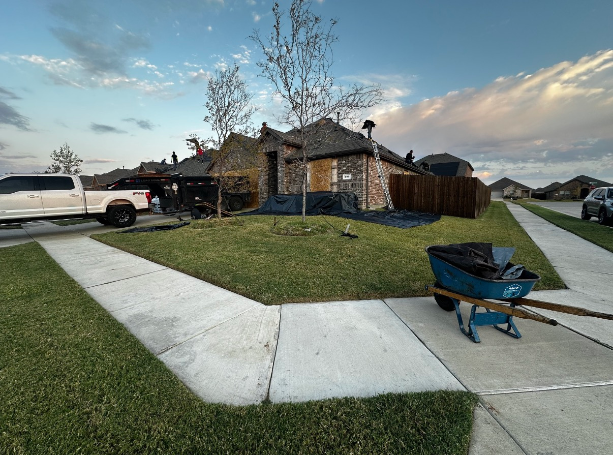 A residential home under roof repair with workers on the roof, a white pickup truck, and a blue wheelbarrow on the sidewalk.