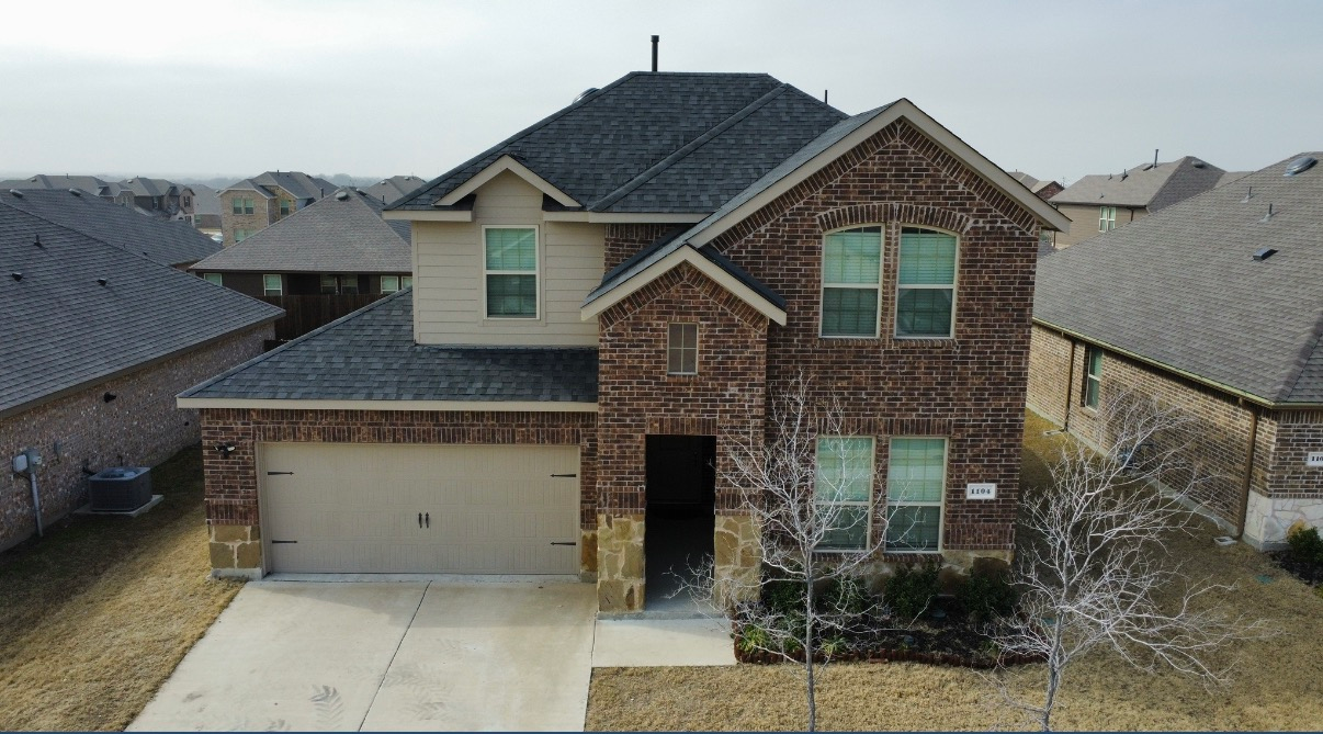 A two-story suburban house with a brown brick facade, gray shingled roof, attached garage, and a front driveway.