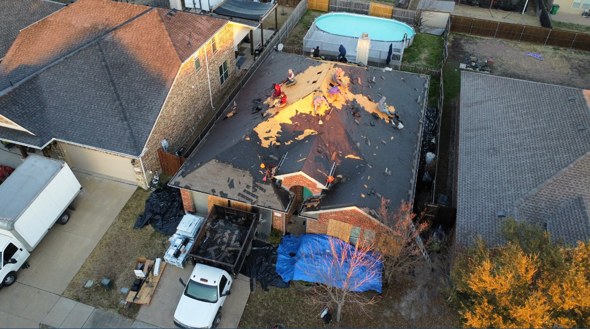 An aerial view shows workers on a residential roof removing shingles, with construction vehicles and debris below.