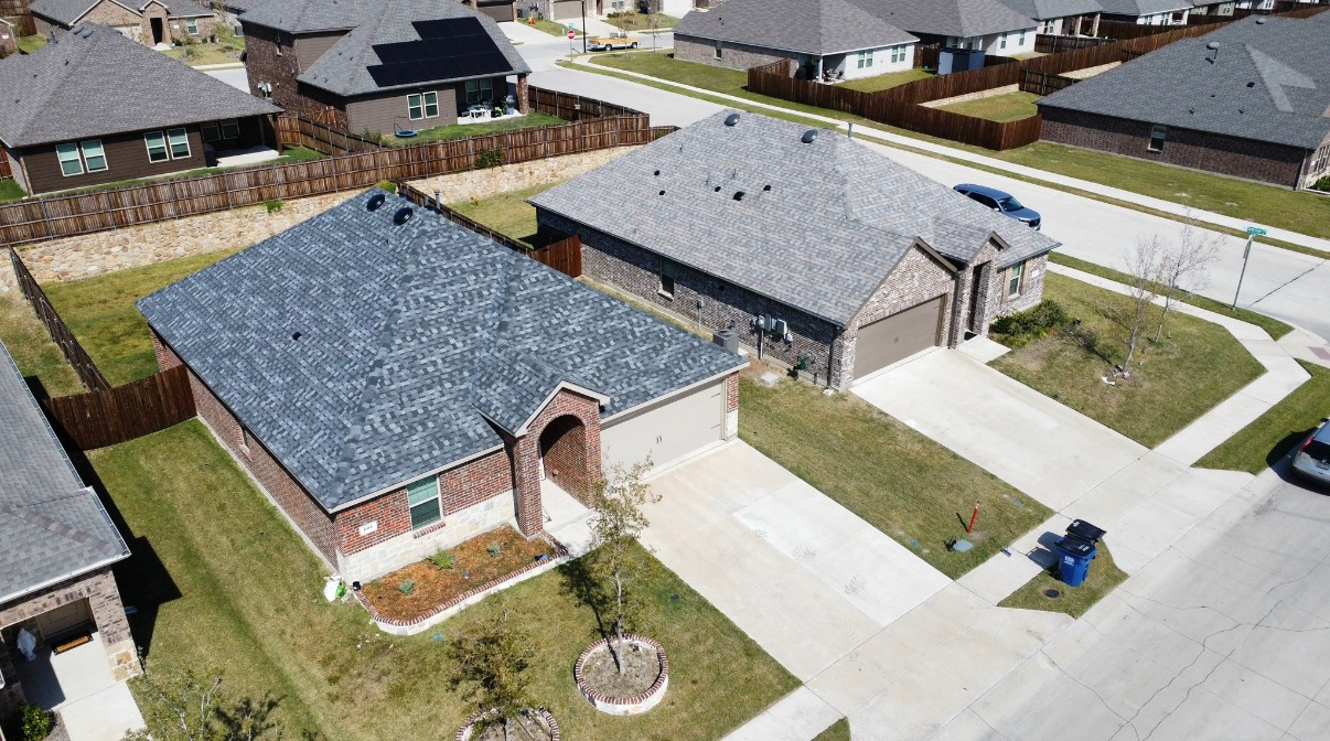 An aerial view of a suburban neighborhood featuring single-story houses with gray shingled roofs and concrete driveways.