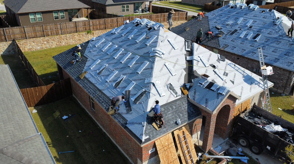 Roofers work on a residential roof covered in silver underlayment during a sunny day.
