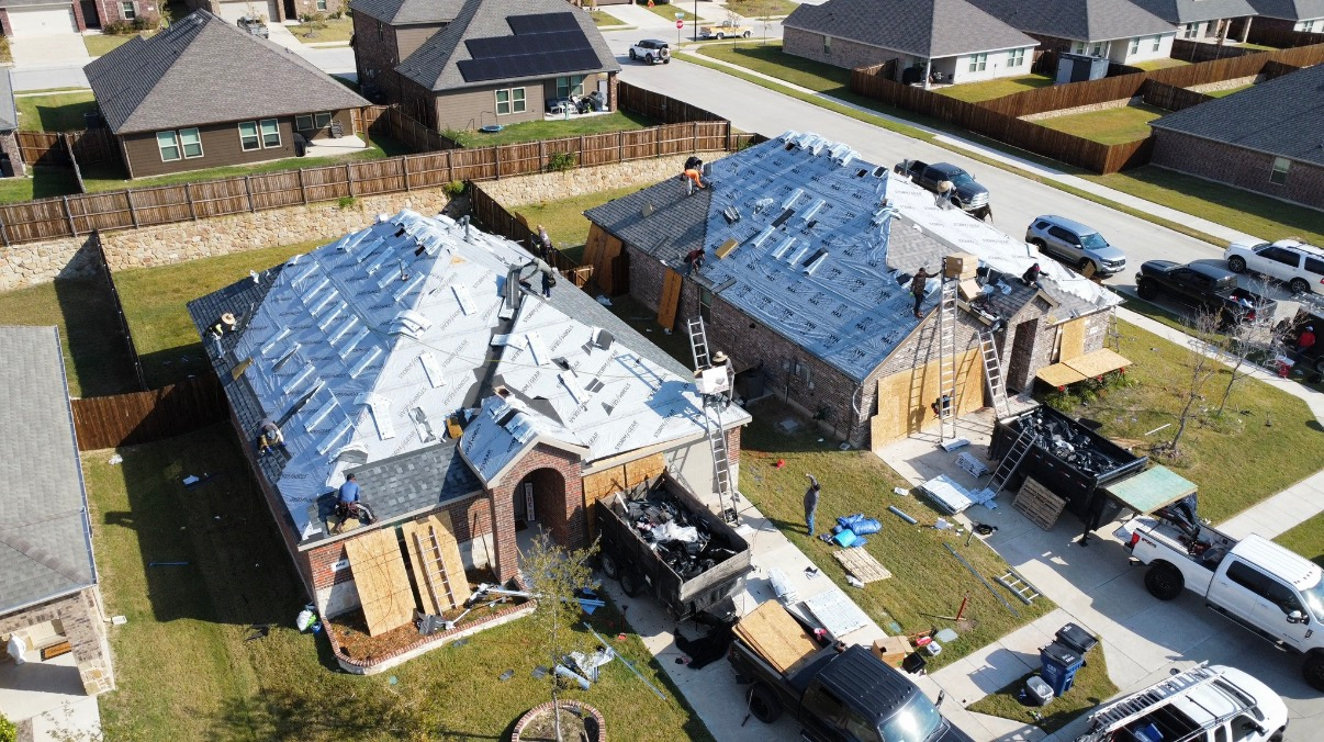 An aerial view of two residential homes undergoing roof repairs, with construction materials and work trucks nearby.