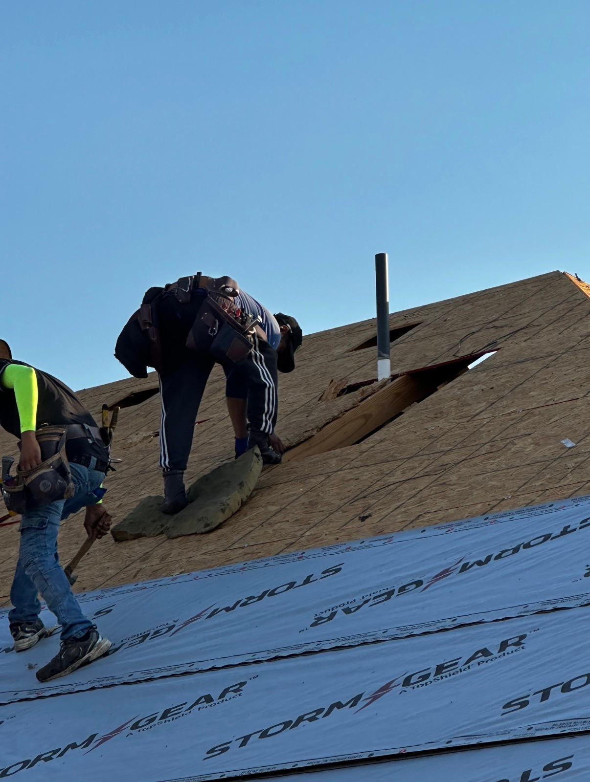 Two people installing roofing materials on a wooden house frame under a clear blue sky.