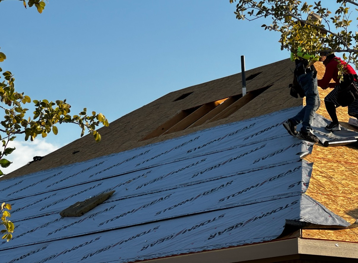 Workers in red and blue clothing install underlayment on a sloping wooden roof under a clear blue sky.