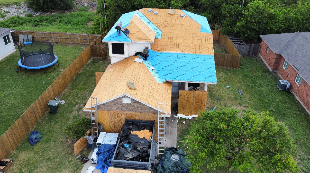 An aerial view of a house undergoing roof replacement, showing bare wooden decking and sections of blue underlayment.