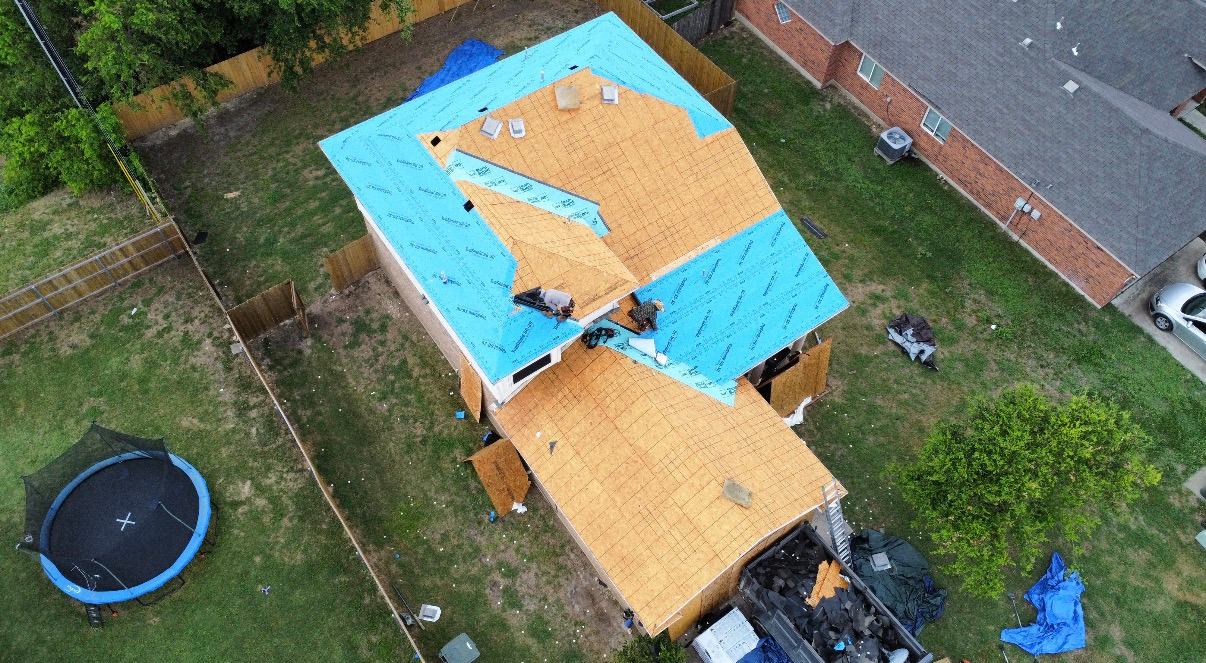 An aerial view of a house undergoing roof repairs, featuring exposed plywood, blue underlayment, and workers on the roof.