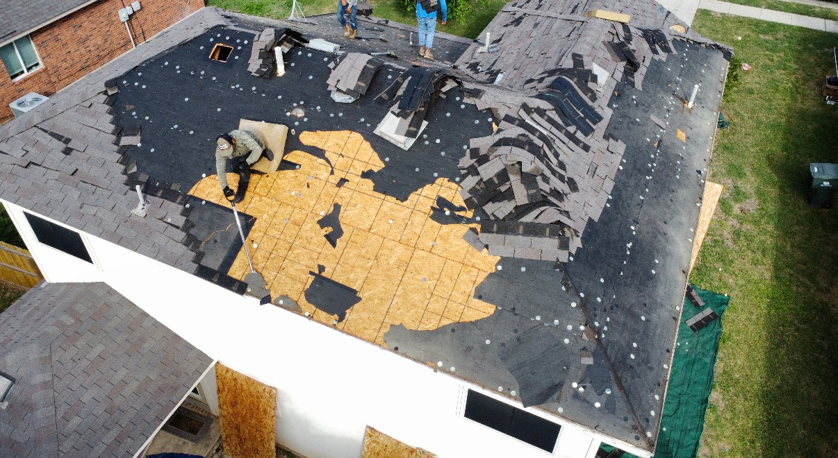 Workers removing old shingles from a residential roof, revealing the wooden decking underneath.