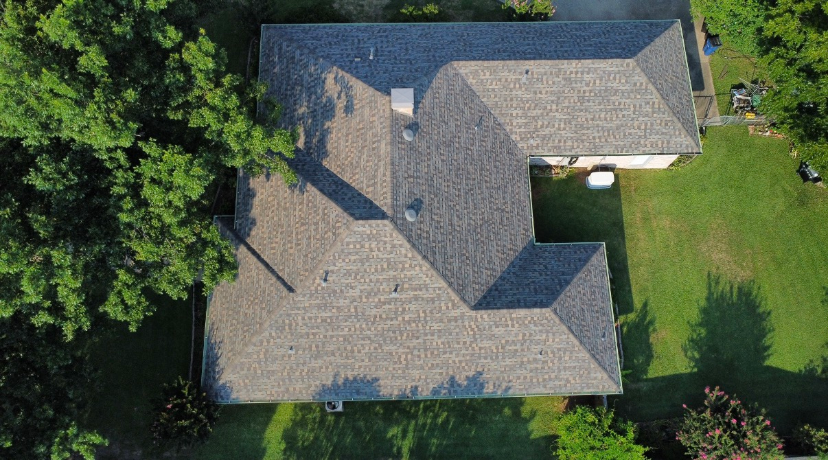 A top-down aerial view of a house with a gray shingled roof, surrounded by green lawns and trees.