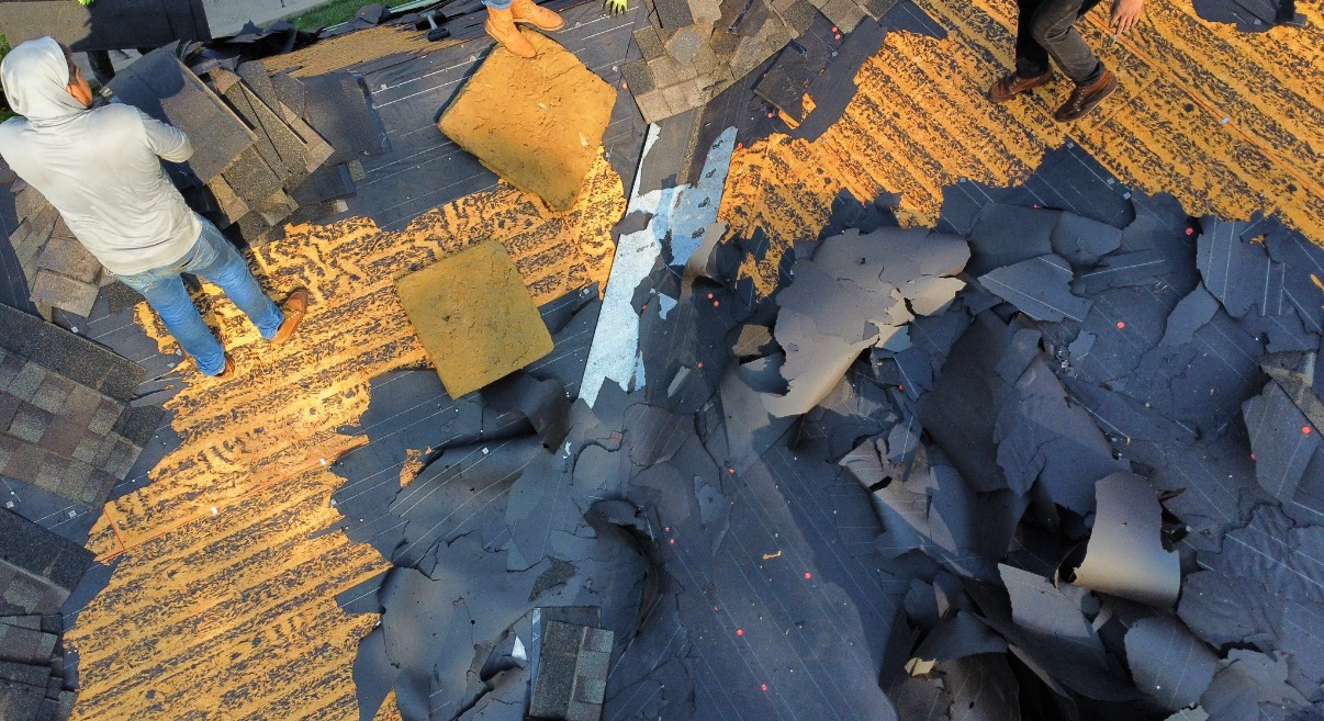 Workers remove old shingles from a wooden residential roof, exposing the underlying deck.