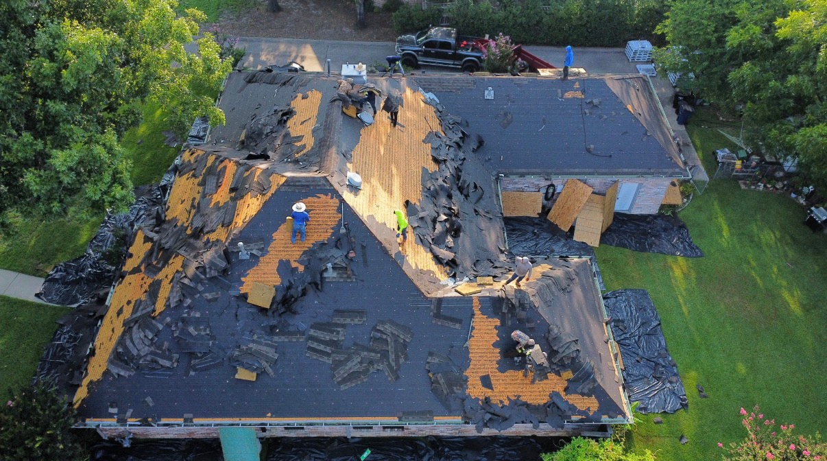 An aerial view of a house undergoing a roof replacement, showing workers removing old shingles to reveal the wood decking.