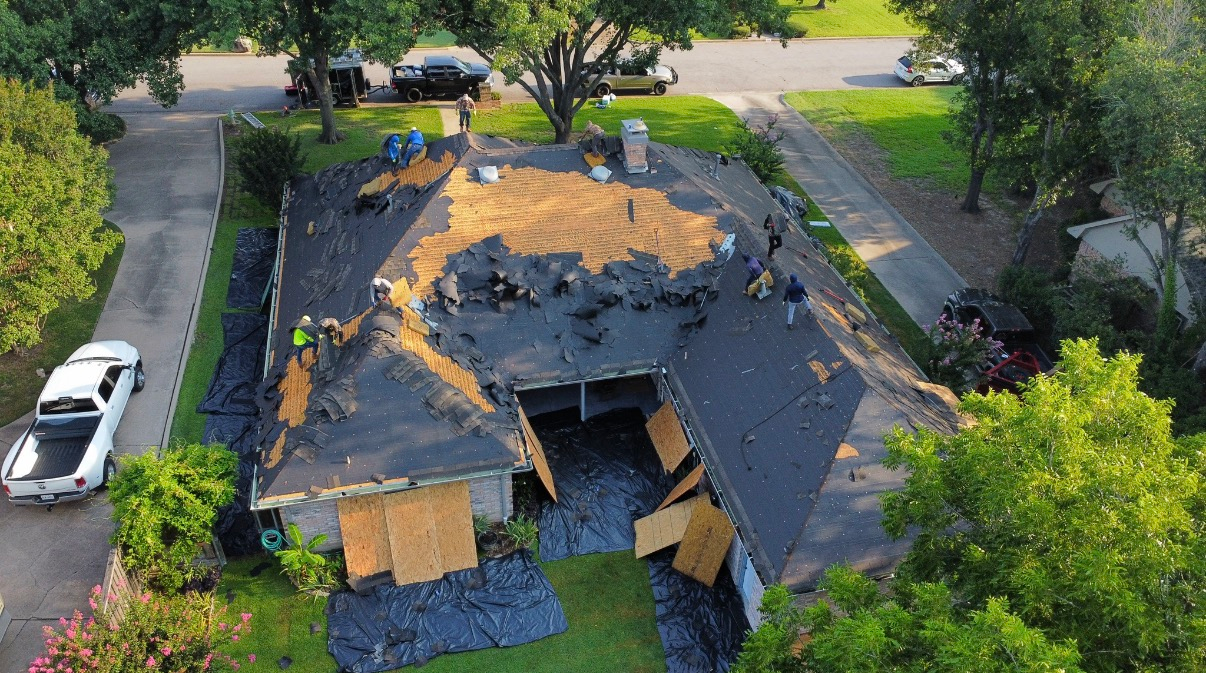 An aerial view of a residential roof replacement in progress, with workers removing old shingles and plywood panels.