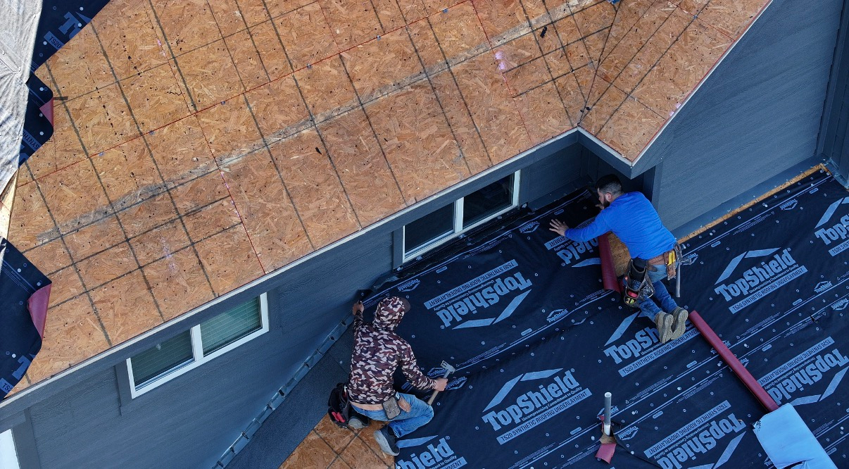 Two roofers install black underlayment on a plywood roof next to a grey house with white-trimmed windows.