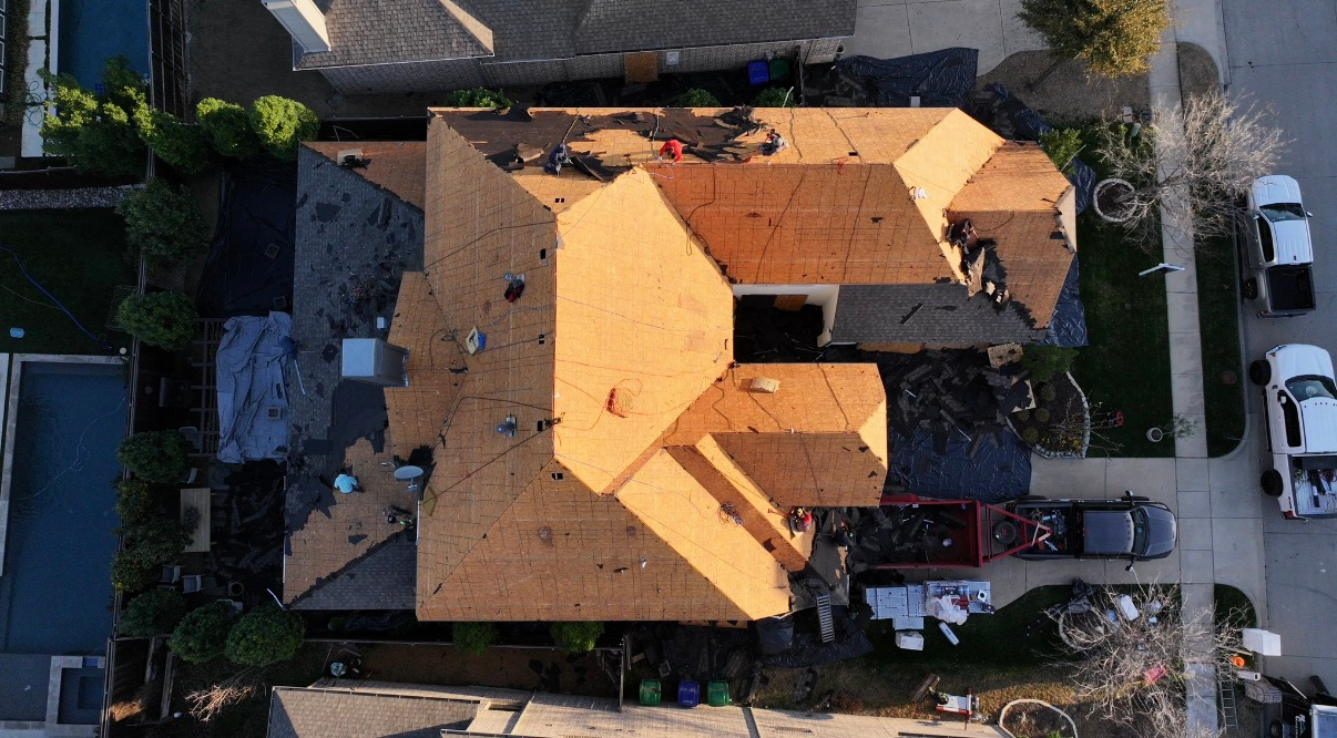 An aerial view of a residential roof undergoing construction, featuring exposed plywood decking and patches of old shingles.