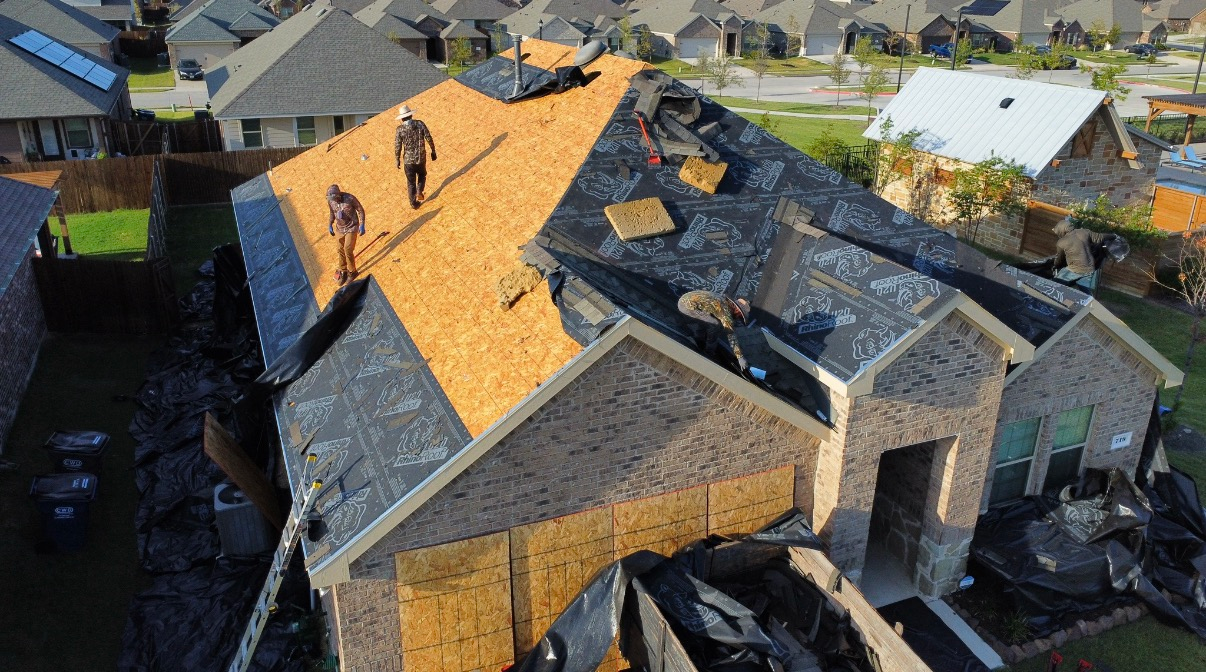 Roofers installing new shingles on a suburban house, with sections of exposed plywood and black roofing felt visible.