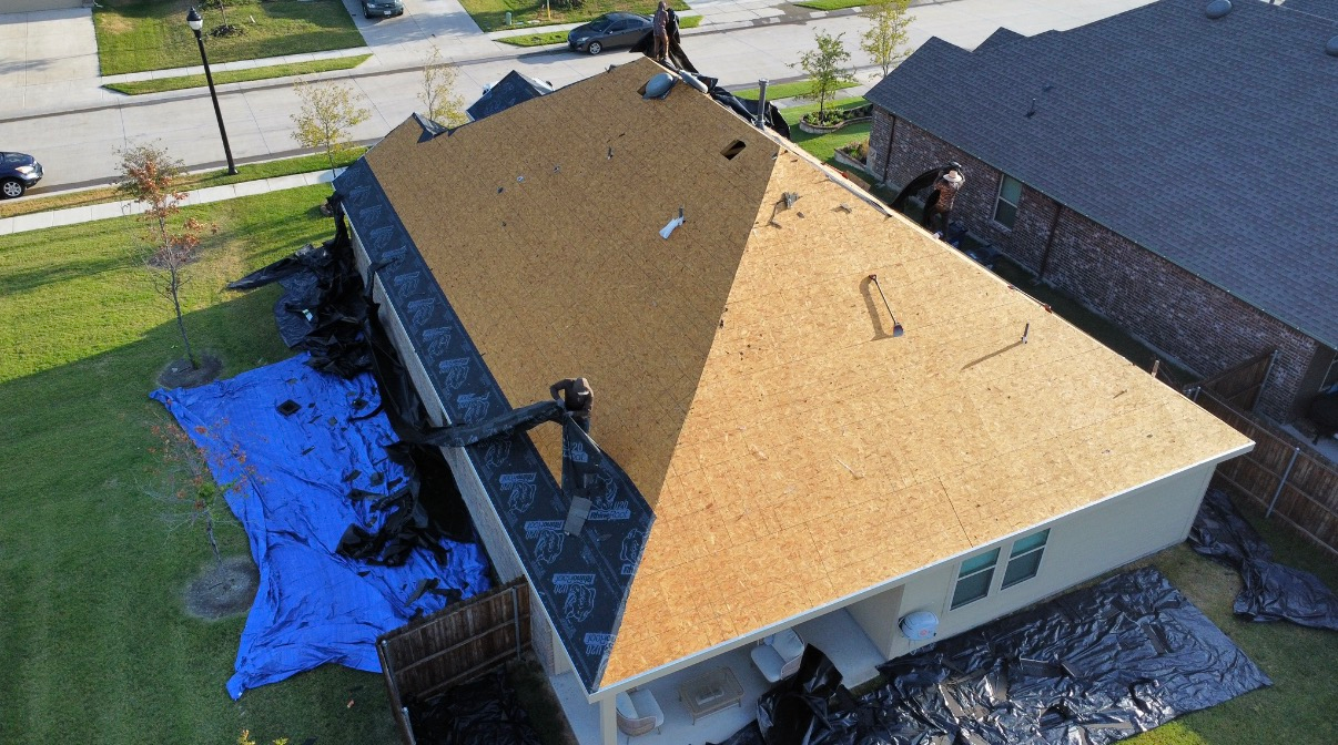 An aerial view of a house roof undergoing repairs, showing exposed wooden decking and a large blue tarp on the ground.