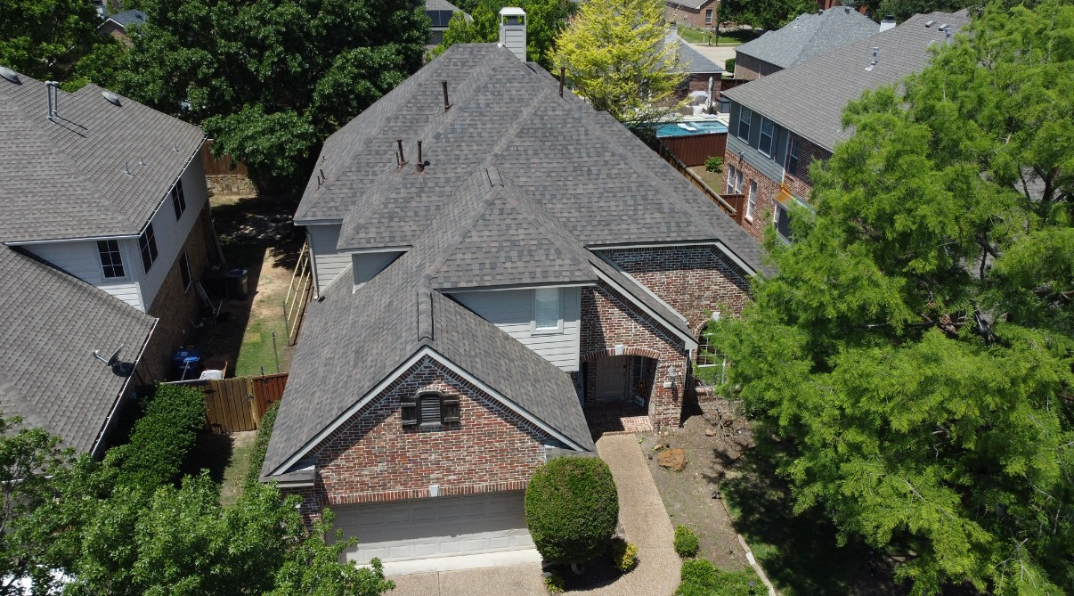 An aerial view of a single-family residential home with a grey roof and brick accents, surrounded by trees and houses.