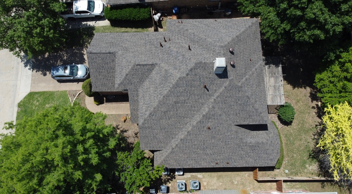An aerial view of a gray shingled residential roof, with a chimney, surrounding trees, a driveway, and two parked cars.