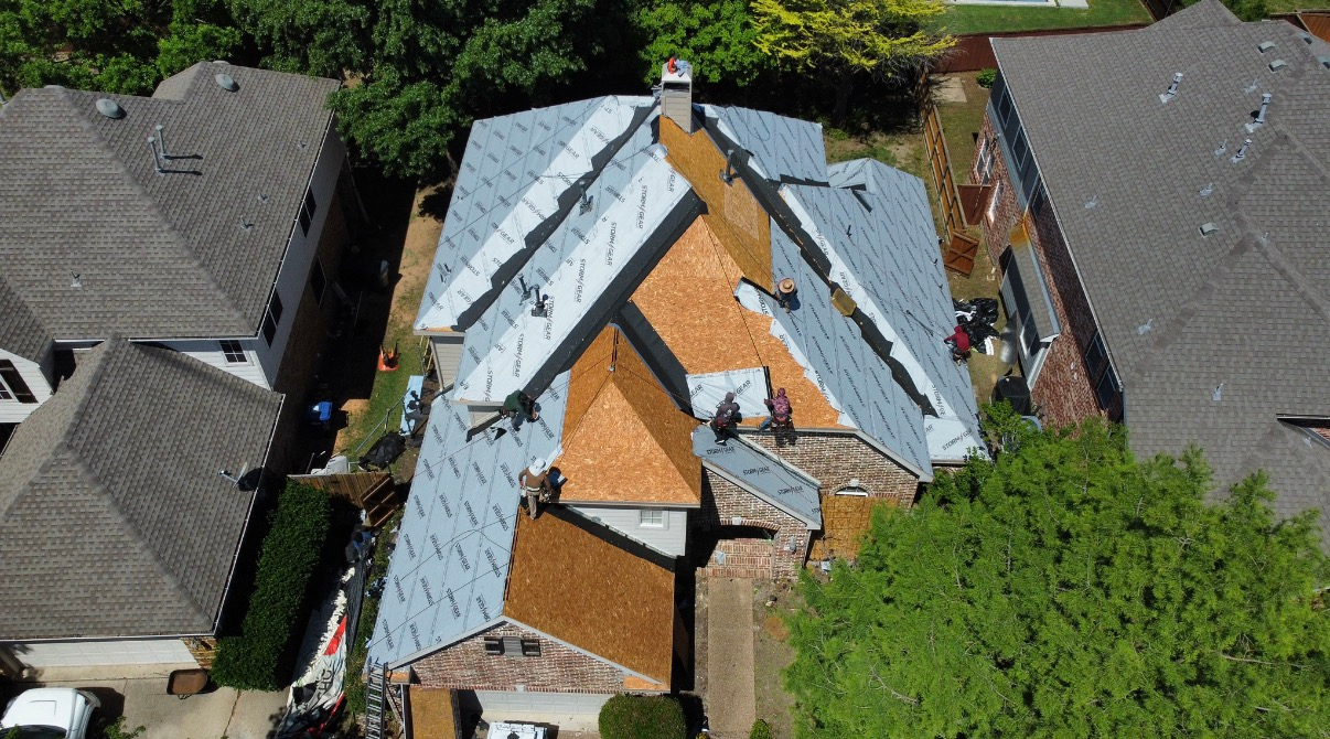 An aerial view shows workers installing new cedar shake shingles on a residential roof partially covered in gray underlayment.
