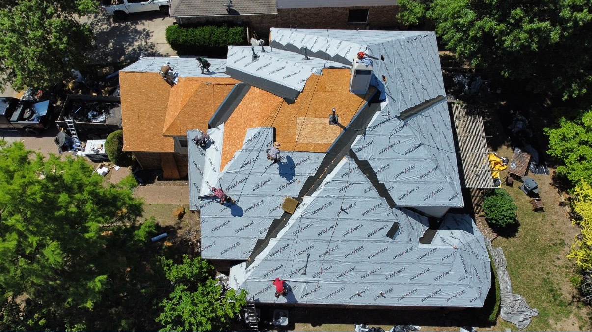 Aerial view of workers replacing a residential roof, showing sections of new orange shingles and gray underlayment.