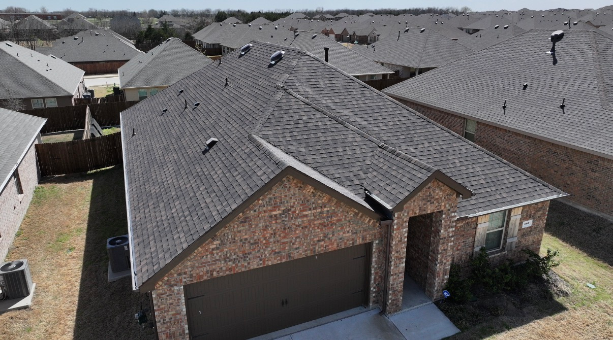 An elevated, high-angle view of a brick house with a dark shingled roof in a suburban neighborhood.