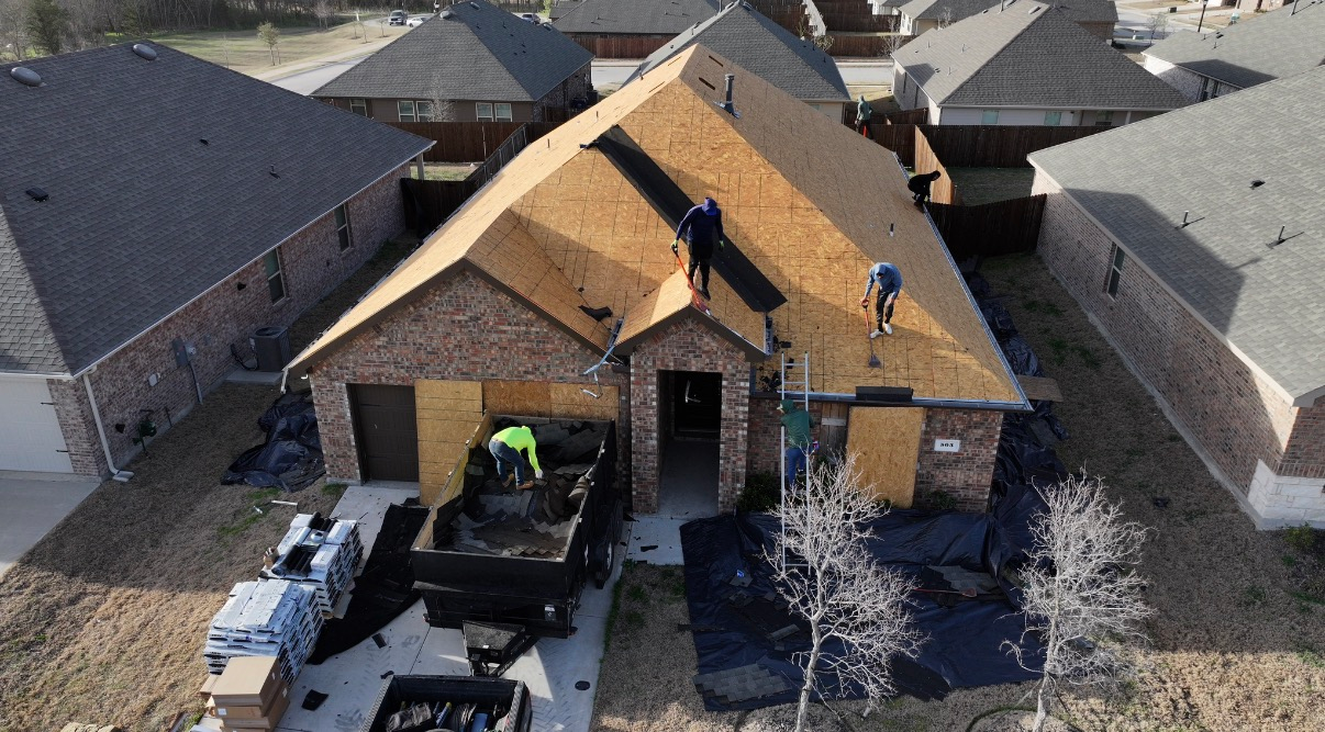 Construction workers replace the roof on a suburban brick home, with a dump trailer parked in the driveway below.