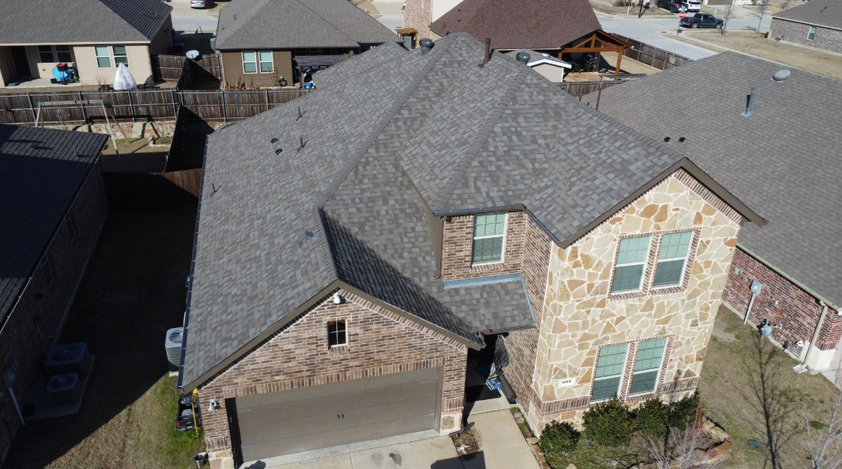 An aerial view of a two-story suburban brick house with a gray shingled roof, a garage, and a small front yard.
