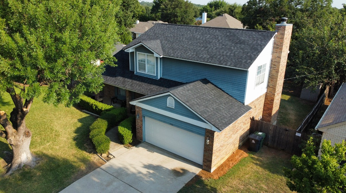 An aerial view of a two-story blue house with a brick chimney and garage, surrounded by green trees and a paved driveway.
