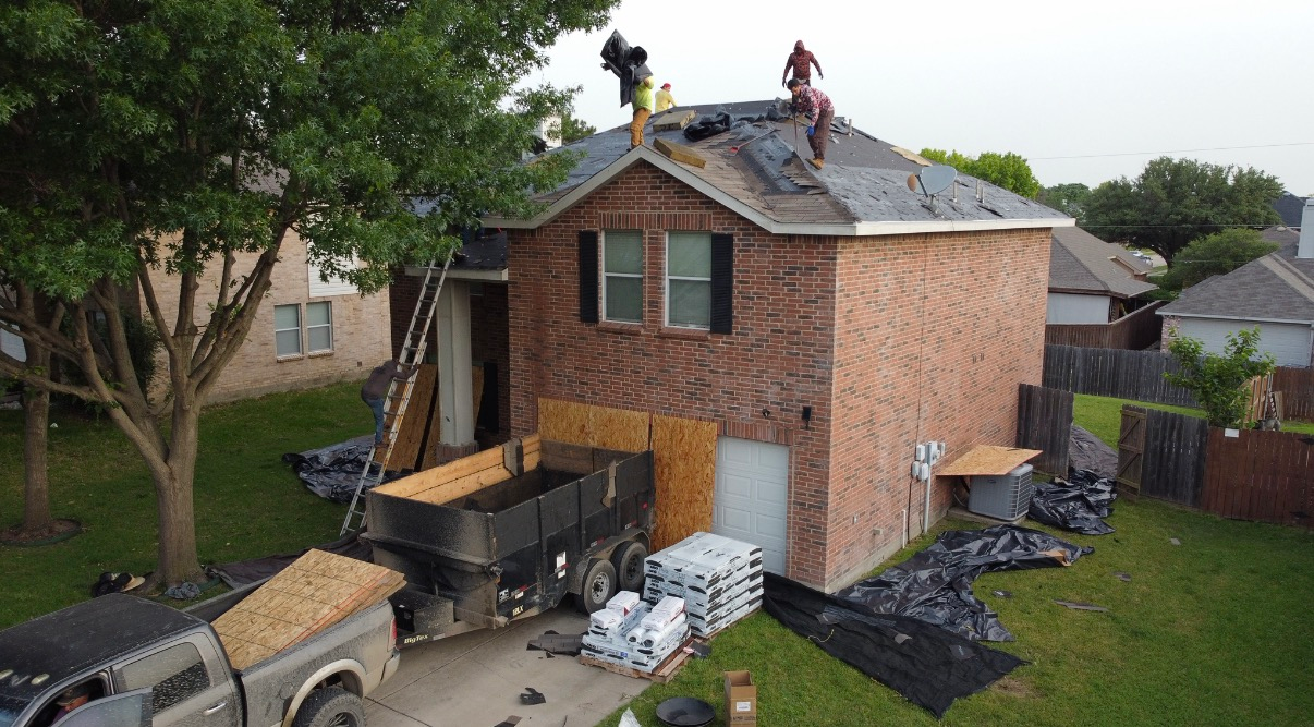 Workers replace shingles on a two-story brick home, with a trailer and supplies parked in the driveway below.