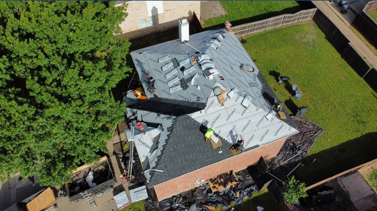 An aerial view of a house roof undergoing construction, featuring grey underlayment, a dark shingled section, and workers.